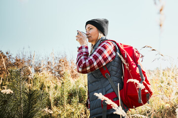 Woman taking break and relaxing with cup of coffee during summer trip. Woman standing on trail and looking away. Female with backpack hiking through tall grass along path in mountains