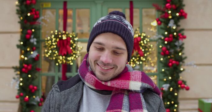 Young caucasian man standing on decorated street, smiling and shaking Christmas gift. Christmas mood concept.