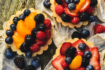 Above tabletop view of four inch tarts with custard filled baked pastry crust shells topped with glazed blueberries, red raspberries, blackberries, sliced strawberries, orange sections