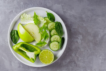 Sliced green fruits and vegetables on a gray background. Fresh green diet food in a white plate. Healthy vegetarian food concept. Copy space. Top view