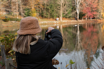 Fototapeta na wymiar A photographer girl taking nature in autumn. Beautiful lakefront view She has brown hat. Hobby concept. Back view. 