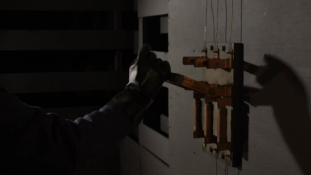 In dramatic lighting a black rubber gloved hand reaches into the frame to grab the wooden handle of an open old large industrial electrical bar switch then hesitates and prepares before shoving it on.