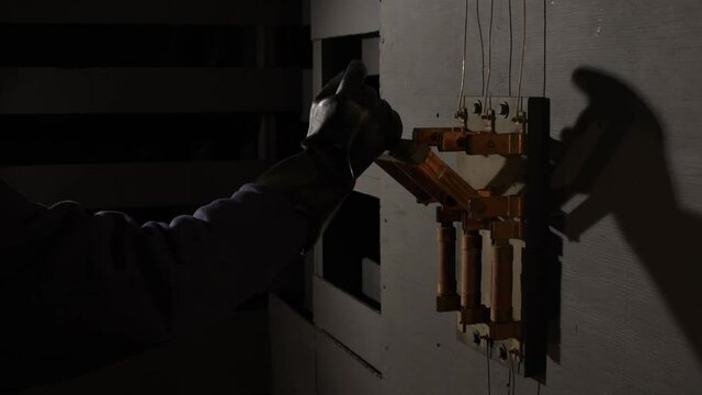 In dramatic lighting a black rubber gloved hand reaches into the frame to grab the wooden handle of an open old large industrial electrical bar switch then shoves the bar to closed.