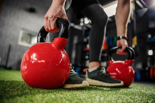 Kettle bells in focus. The masculine strong arms take the sports equipment for exercising in both arms and prepare to lift weights. Two red kettle bells on green artificial grass indoor modern gym