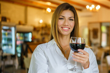 Front view portrait of beautiful happy brunette caucasian woman smiling while holding a glass of red wine looking to the camera standing at restaurant in day wearing white shirt copy space