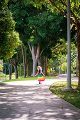 A little girl in a rainbow dress fun running on the concrete path while looking back in the park. Sunny day dapple lights.