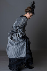 A Victorian woman wearing a striped silk polonaise and black skirt alone against a studio backdrop