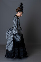 A Victorian woman wearing a striped silk polonaise and black skirt alone against a studio backdrop