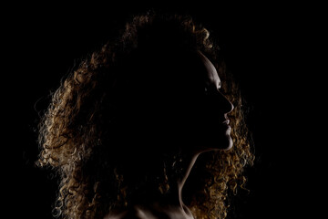 Side lit brunette girl with long curly hair. Silhouette studio portrait on dark background..