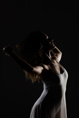 Girl with curly hair making ballet poses. Side lit silhouette of ballerina in white dress against black background.