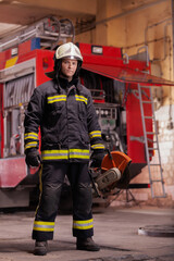 Professional firefighter in uniform wearing protective helmet and standing with emergency rescue equipment. Circular hydraulic cutting hard tool. Firetruck in the background.