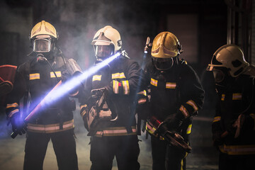Naklejka premium Group of professional firefighters wearing full equipment, oxygen masks, and emergency rescue tools, circular hydraulic and gas saw, axe, and sledge hammer. smoke and fire trucks in the background.