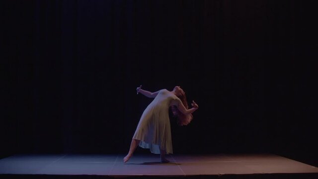 A female dancer is dancing on a stage, there is a blue and orange light flickering above her. Slow motion