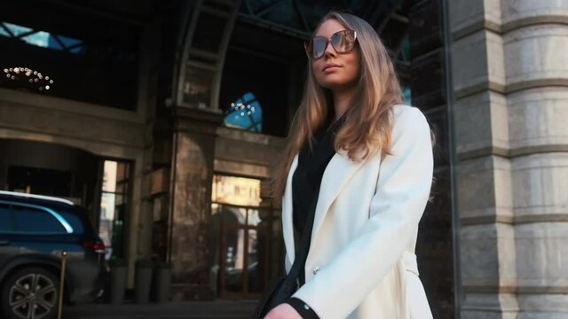 A girl in glasses and a white coat walks through the business district of the city. Against the backdrop of luxury cars and apartments