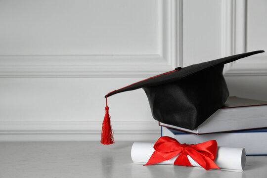 Graduation Hat, Books And Diploma On Floor Near White Wall, Space For Text