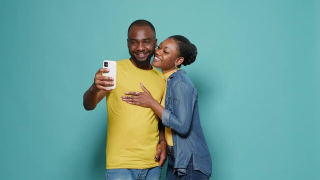 Couple taking selfies with smartphone in front of studio camera. Man and woman making silly faecs while they take pictures together on mobile phone. Casual people enjoying leisure activity