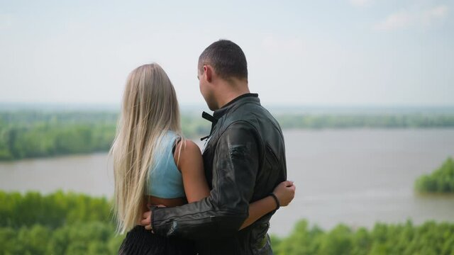 Couple of long haired woman and professional biker embraces standing on hill against picturesque lake on spring day backside view slow motion