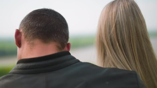 Guy in black leather clothes hugs long fair haired lady standing against picturesque lake on summer day close backside view slow motion