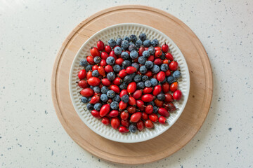 Top view of a white plate with rosehip and blackthorn on a round wooden board