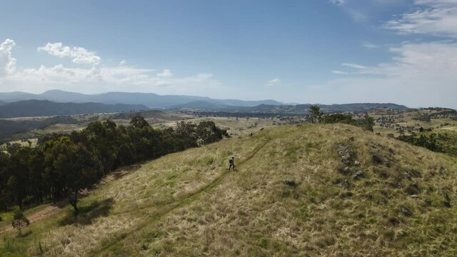 Aerial drone shot looking down on the small figure of a man walking up a steep grassy hill surrounded by trees. In the background you can see blue mountains and cloudy sky on the horizon. 