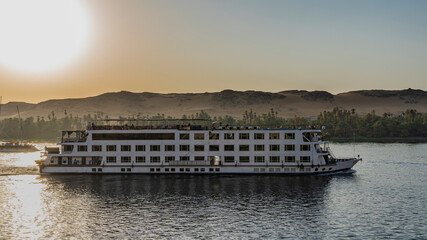 Fototapeta premium A white cruise ship is sailing on the Nile River. The evening sun shines over the coastal sand dunes. Reflection on the water. Egypt
