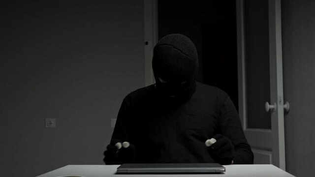 Burglar under disguise steals money and jewelry rummaging in drawer and takes laptop from table surface sitting at wooden desk in flat closeup.