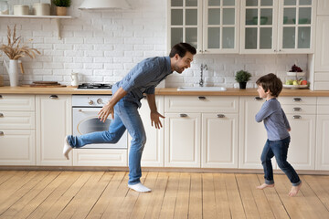 Happy Caucasian father with little son dancing in kitchen at home, smiling young dad and cute 5s boy child having fun, moving to favorite music, engaged in funny activity, spending leisure time