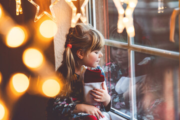 Little preschool girl holding cup Santa Claus boot with gift called Nikolausstiefel in German. Happy child wait on holiday by window with Christmas lights in winter. Cozy family celebration of xmas.