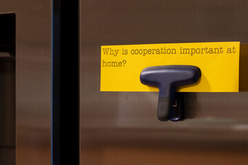 Closeup of a note attached to the refrigerator door with a magnetic clip, which says - why is cooperation important at home? Kids education concept - cooperation starts at home.