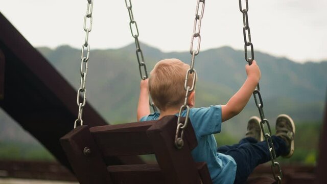 Toddler boy plays handmade swings holding on to chains on wooden playground against large mountain silhouettes close backside view slow motion
