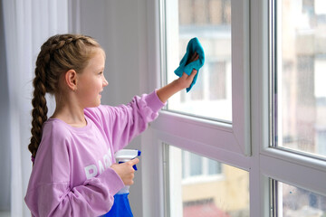 A little girl in a purple sweater sprinkles water from a bottle on the window and wipes it off with a rag. The child helps with cleaning the house. Clean the window.