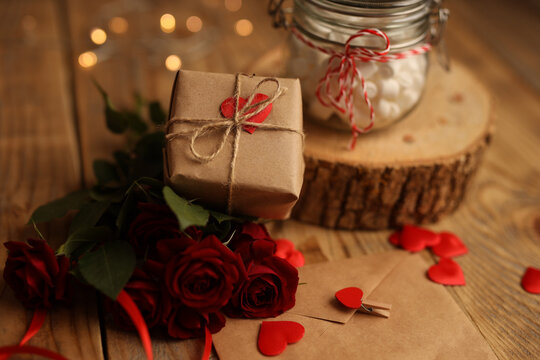 A gift in craft paper on a wooden background, red roses around, a jar of marshmallows, a letter in an envelope and hearts. Picture for Valentine's Day. The concept of gifts and lovers. High-quality ph