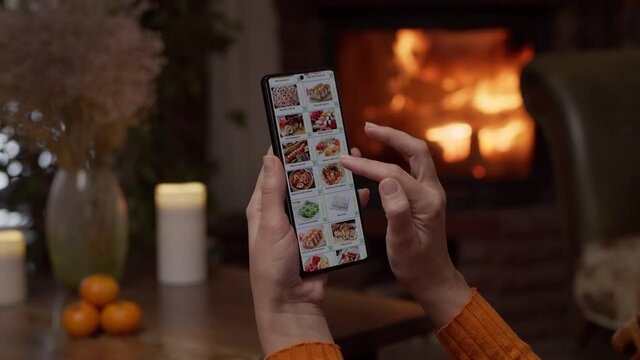 Ordering food using a smartphone at home. A woman selects sushi sets in the internet menu of a Japanese restaurant using an application on a smartphone. Home evening furnishings with a burning fire in