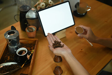 Photo of young barista man sitting in front of the white blank screen digital tablet on the wooden table surrounded by coffee equipment.