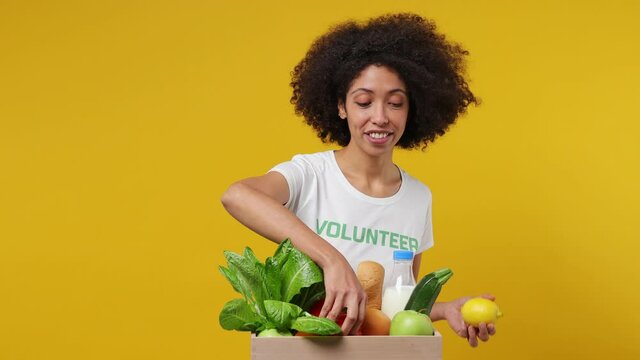 Young black woman 20s wears white t-shirt green title volunteer put fruit vegetable in basket isolated on plain yellow background studio portrait. Voluntary free work assistance help charity concept