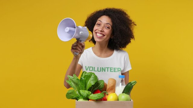 Vivid young black woman 20s in white t-shirt green title volunteer with fruit vegetable basket scream in megaphone isolated on plain yellow background studio portrait. Voluntary free work help concept