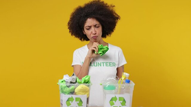 Young black woman 20s in white t-shirt green title volunteer hold rubbish think choose waste bin to recycle isolated plain yellow background studio portrait. Voluntary free work help charity concept