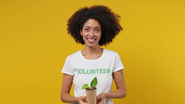 Young black woman 20s in white t-shirt green title volunteer hold in hand give you paper cup with plant isolated on plain yellow background studio portrait. Voluntary free work help charity concept