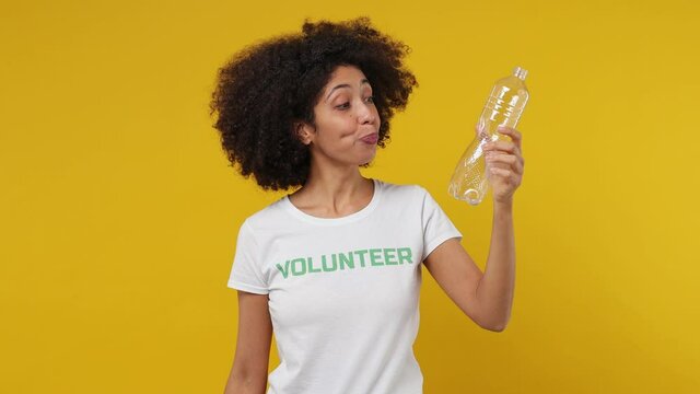 Young black woman 20s in white t-shirt green title volunteer drink water from plastic bottle put in proper waste bin isolated on plain yellow background studio portrait. Stop nature garbage concept