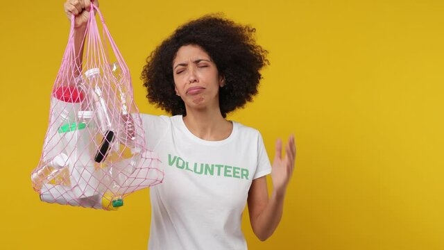 Upset sad fatigued young black woman 20s in white t-shirt green title volunteer hold in hand bag with plastic rubbish isolated on plain yellow background studio portrait. . Stop nature garbage concept