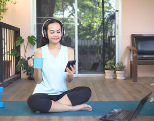 Asian woman wearing headphones, holding coffee cup and reading meassage on smartphone, sitting on...