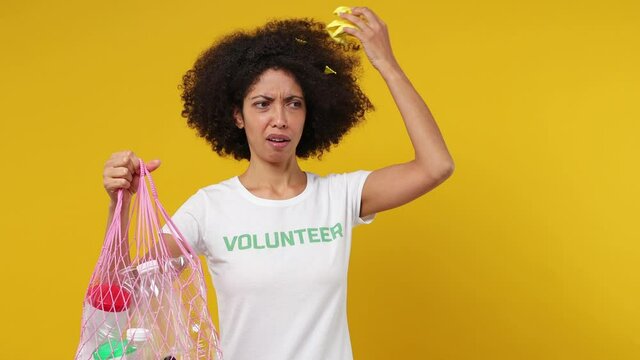 Shocked young black woman 20s in white t-shirt green title volunteer hold bag with plastic rubbish find trash in hair isolated on plain yellow background studio portrait. . Stop nature garbage concept