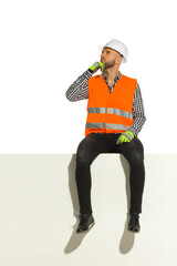 Man in white hardhat and reflective waistcoat is sitting on a top of white banner, looking away and...