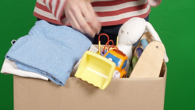 Woman preparing clothes and toys for charity donation. Female hands sorting and packing kid toys, children clothes into cardboard box over green screen chroma key background