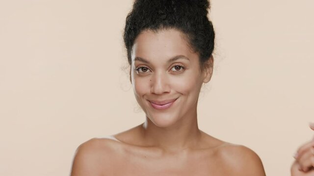 Gorgeous African American young slim woman with dark curly hair and nude shoulders presses her cheek and smiles wide for the camera on beige background | Sagging face skin prevention concept