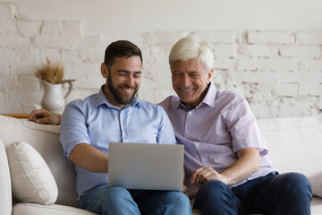 Smiling young man teaching happy middle aged elderly retired father using laptop software applications, sitting on couch at home, enjoying web surfing information or shopping in internet store.