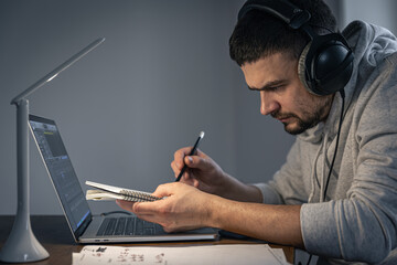 A man in headphones at night in front of a laptop screen writes music.