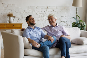 Fototapeta Happy relaxed young man laughing, enjoying pleasant conversation with older retired father, resting together on cozy couch. Joyful carefree two generations family discussing life news at home. obraz