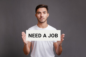 Handsome caucasian man holding sheet of paper with text need a job while standing isolated over gray background, looking for a work, job search concept, toned image