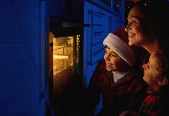 Cheerful happy mom with her adorable curious kids in a red and green plaid shirt sitting on the floor and looking through the oven at the traditional German Stollen bread baked on a baking tray
